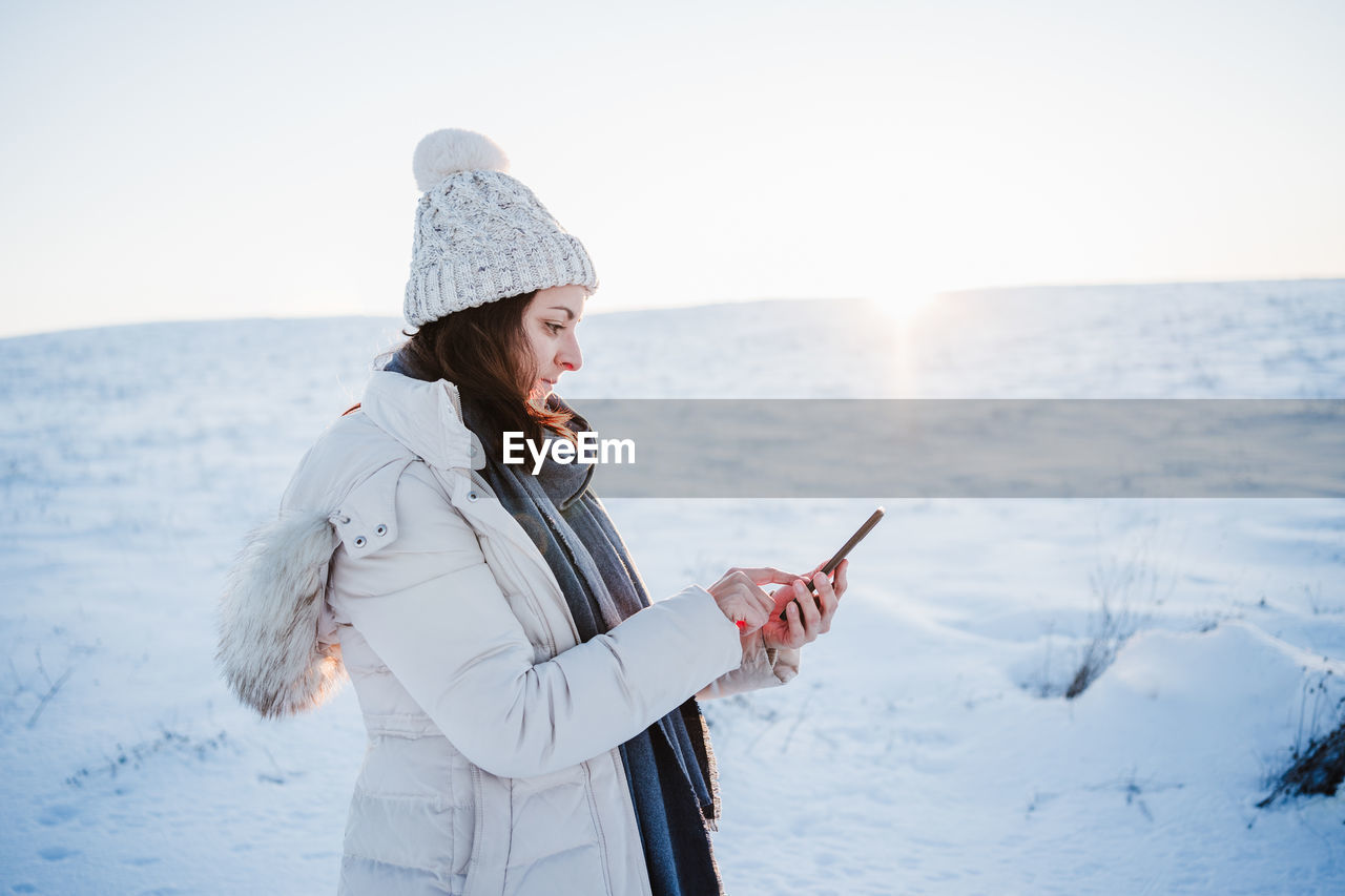 Side view of woman using mobile phone while standing on snow covered land