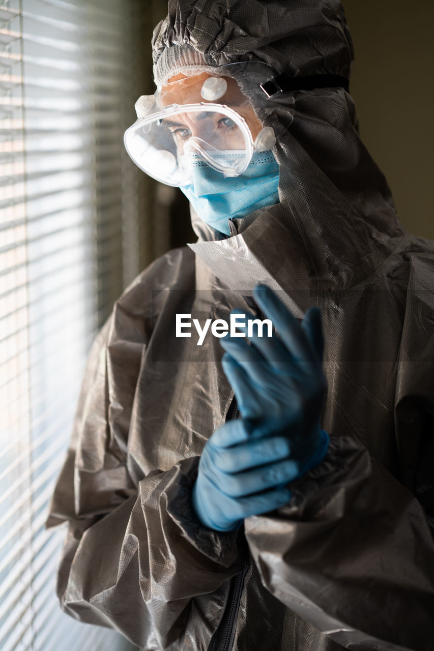 Woman wearing surgical mask and protective suit standing by window