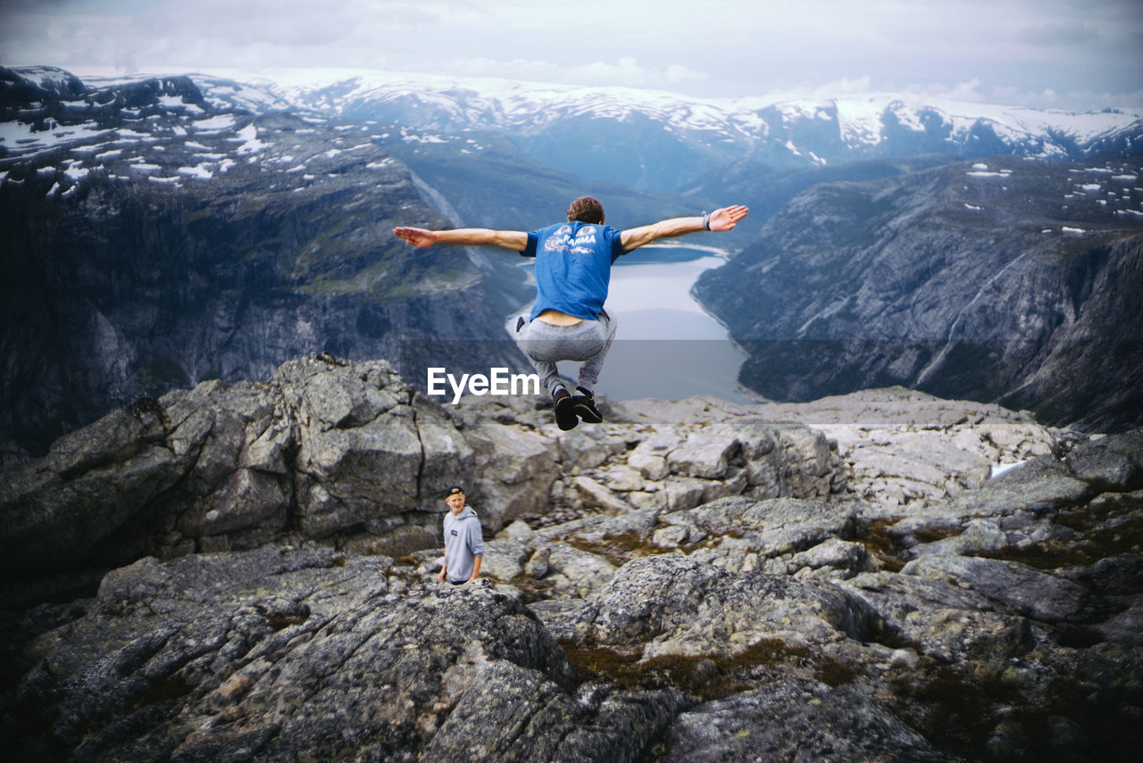 REAR VIEW OF MAN ON ROCK AGAINST MOUNTAIN