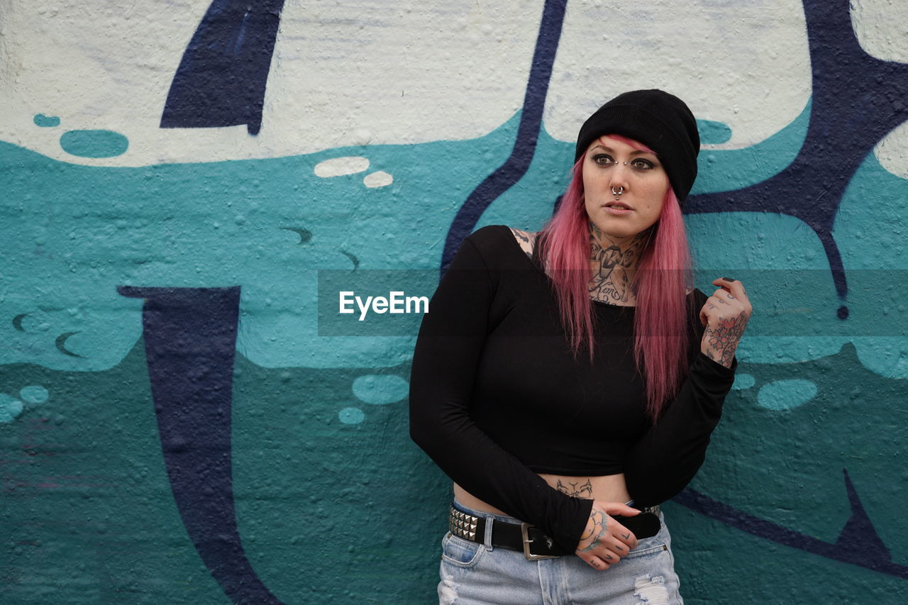 Young tattooed woman with pink hair standing against graffiti wall