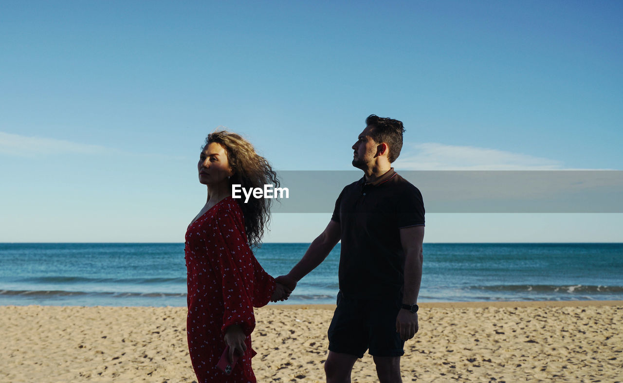 Young woman with man standing at beach against clear sky with holding hands