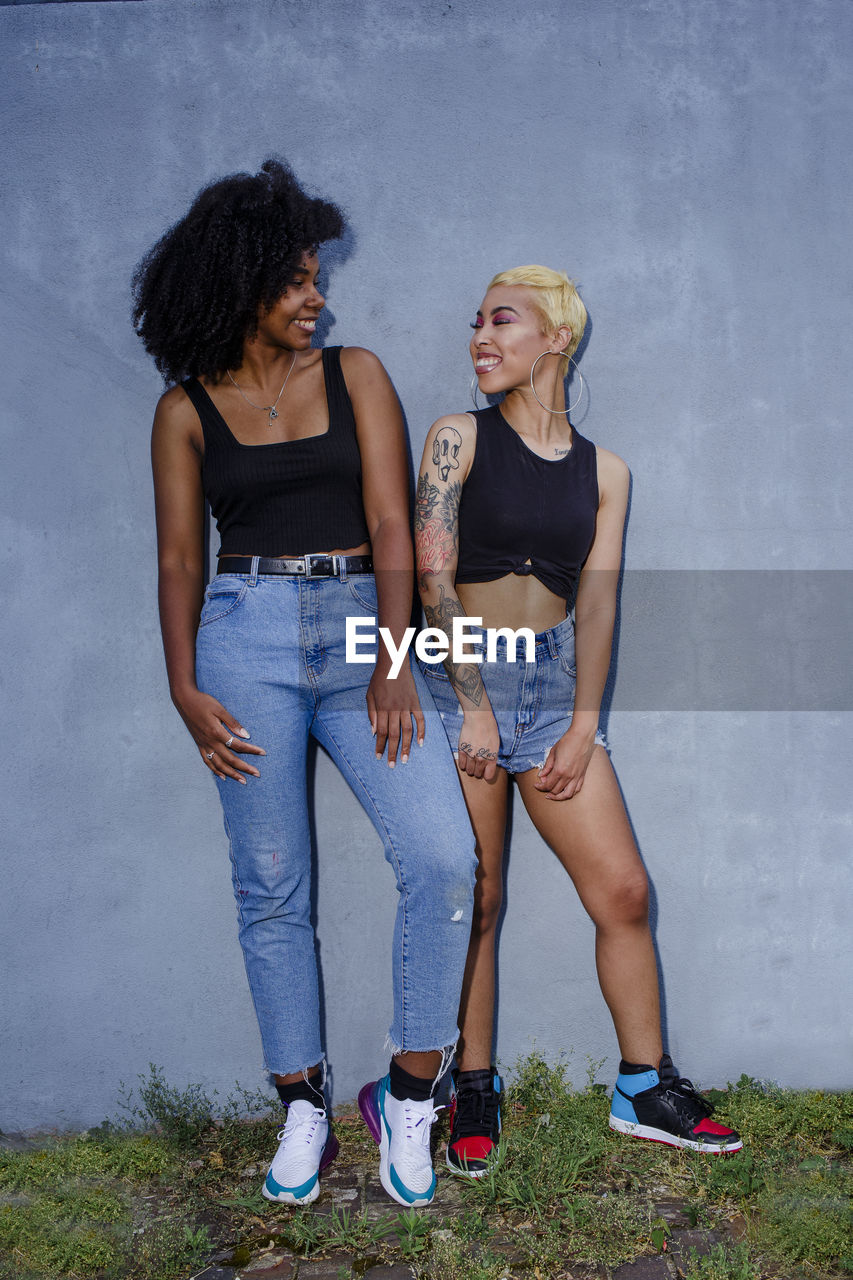 Two smiling friends lean together against a wall in matching clothes