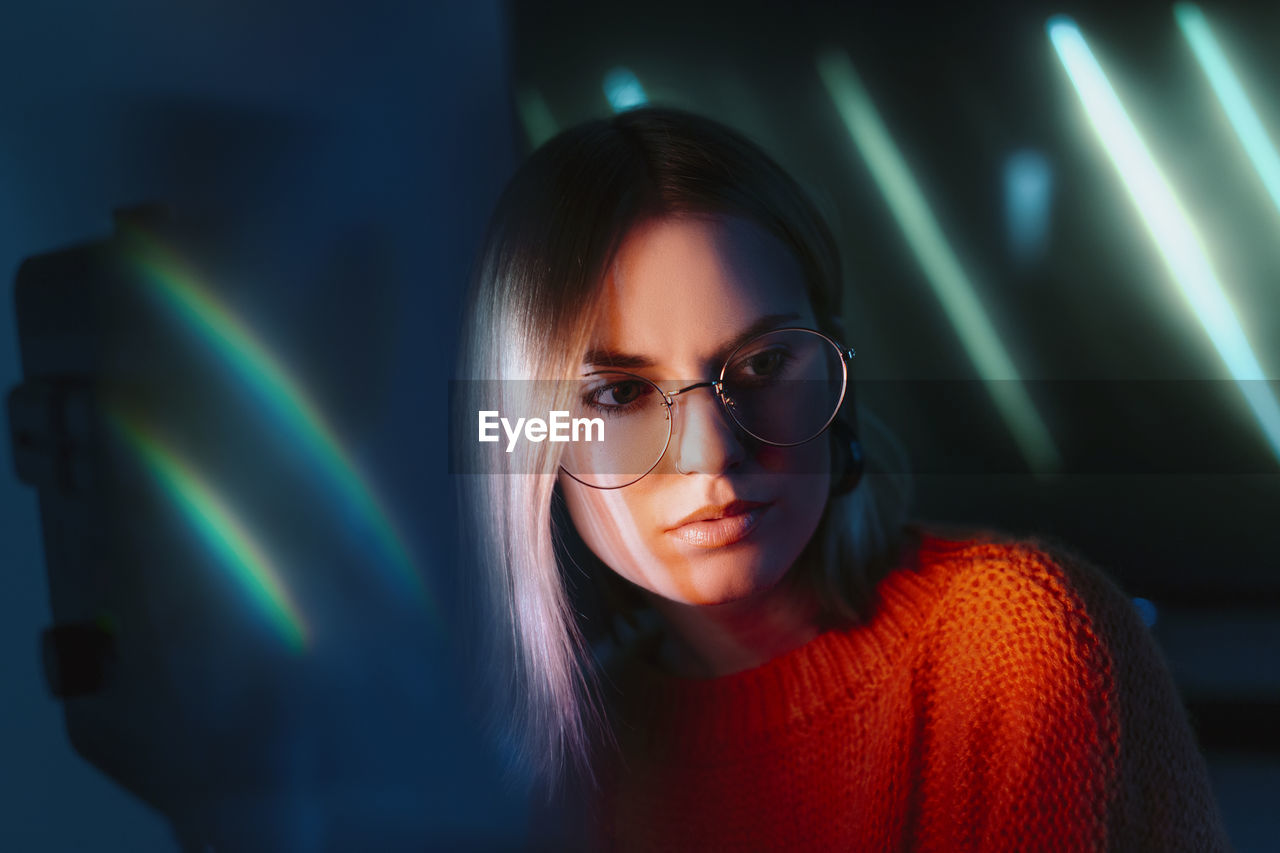 PORTRAIT OF YOUNG WOMAN WITH EYEGLASSES IN ILLUMINATED ROOM
