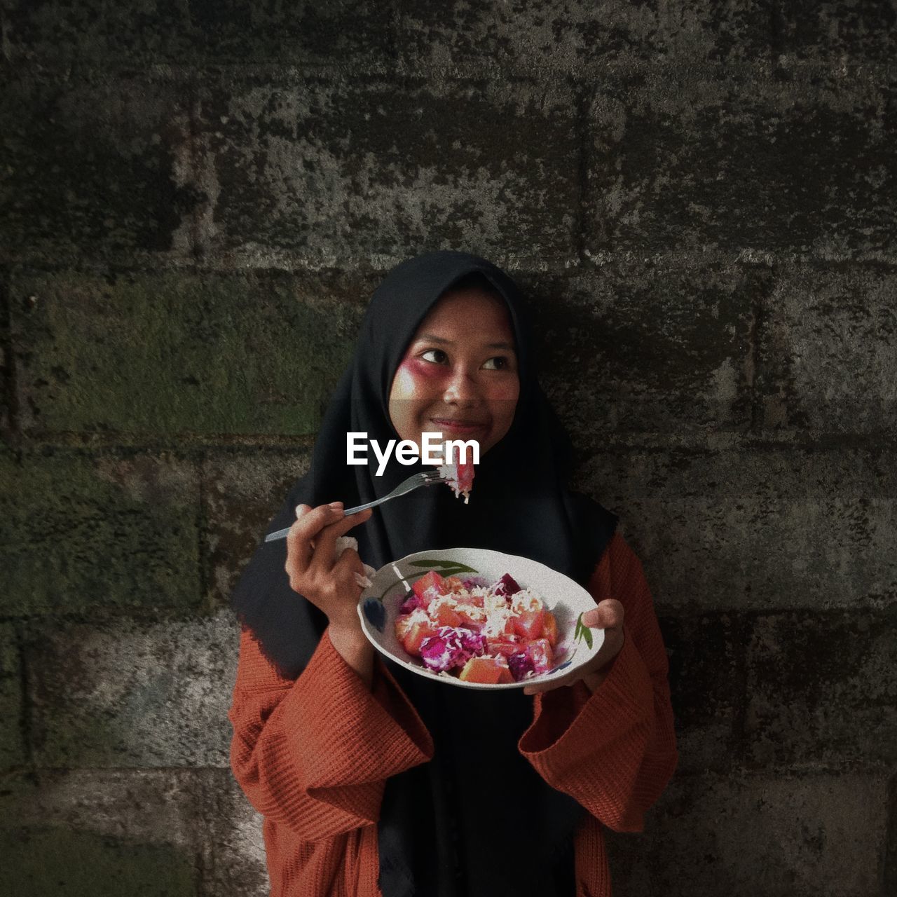 Woman wearing hijab eating food against wall