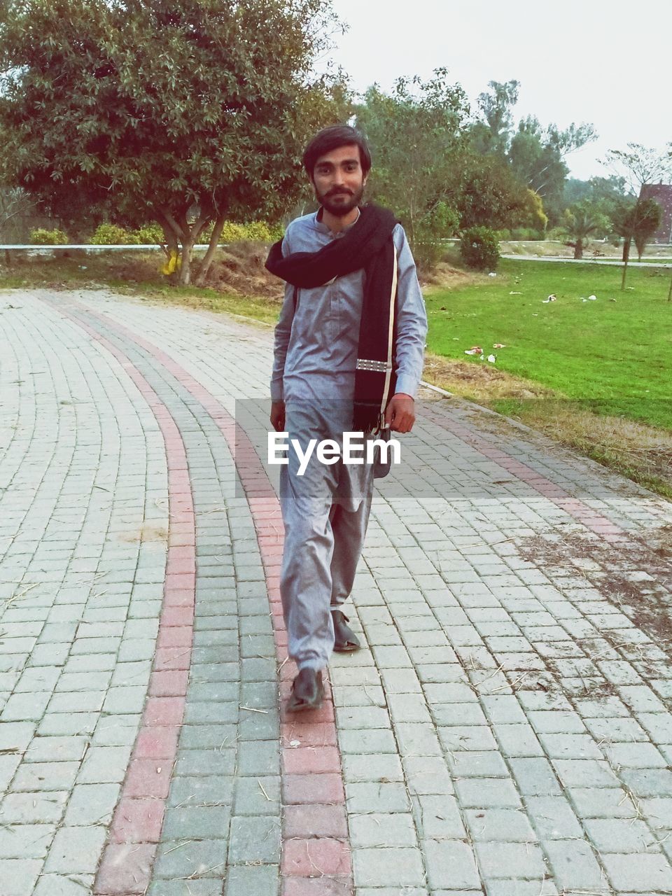 one person, full length, footpath, casual clothing, lifestyles, leisure activity, young adult, day, adult, front view, looking at camera, men, portrait, plant, nature, tree, walkway, outdoors, smiling, standing, clothing, sports, city, street, person, walking, footwear, happiness, emotion, architecture, paving stone, fashion