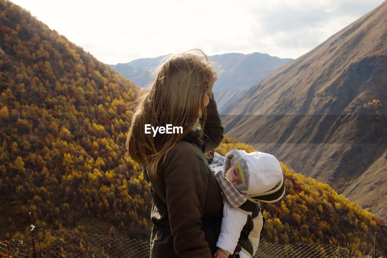 Portrait of woman with her little daughter in ergo carrier on a background of autumn mountains.