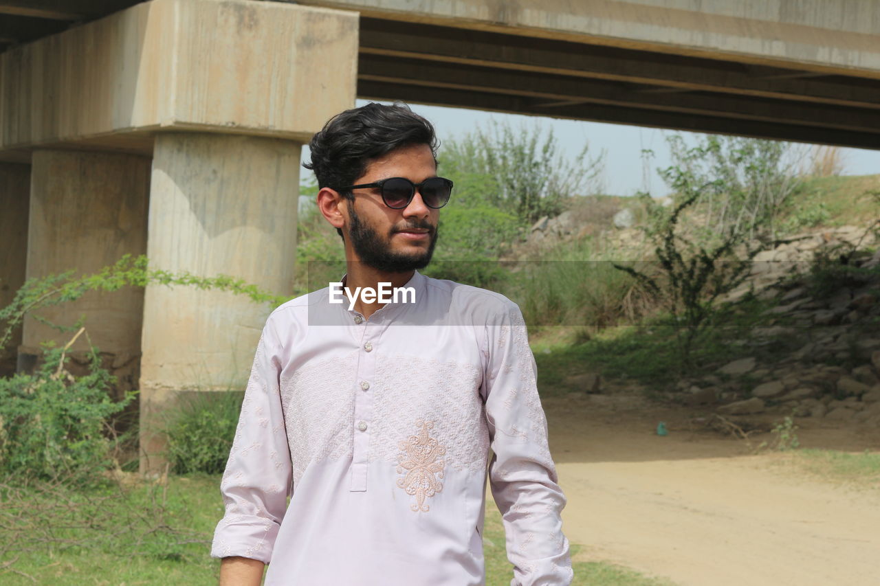 one person, sunglasses, fashion, glasses, young adult, front view, adult, standing, beard, casual clothing, men, shirt, waist up, facial hair, day, lifestyles, architecture, spring, nature, portrait, leisure activity, person, clothing, outdoors, built structure, button down shirt, plant, cool attitude, looking, looking away, three quarter length