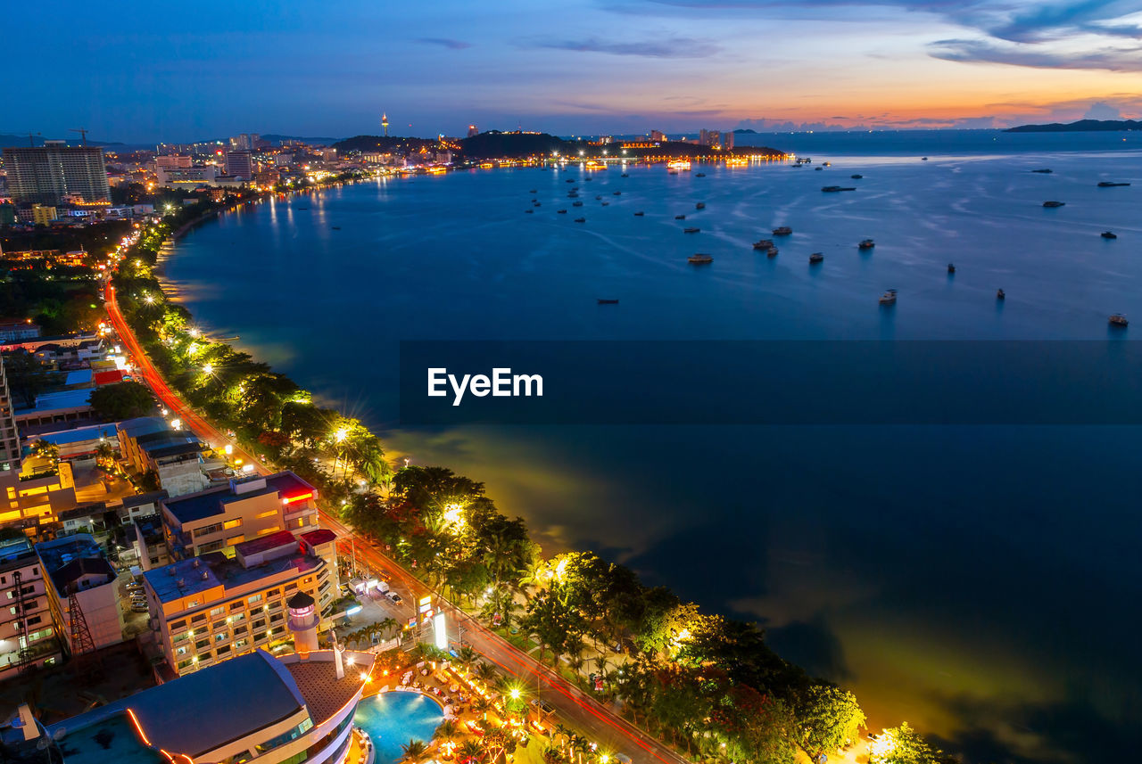 HIGH ANGLE VIEW OF ILLUMINATED CITY BY SEA AGAINST SKY AT SUNSET