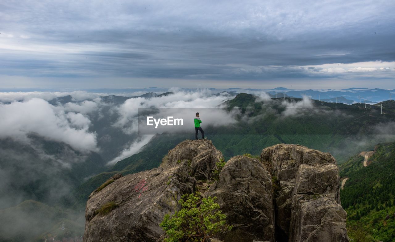 Man standing on cliff overlooking mountains and wind turbine