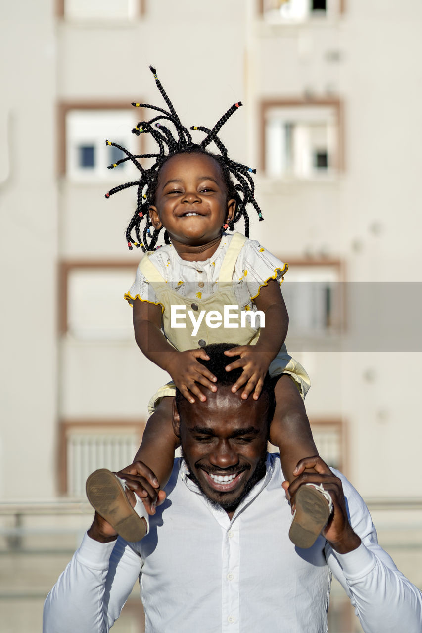Cheerful african american father in shirt carrying little daughter on shoulders and jumping while spending time together on street in city in sunlight