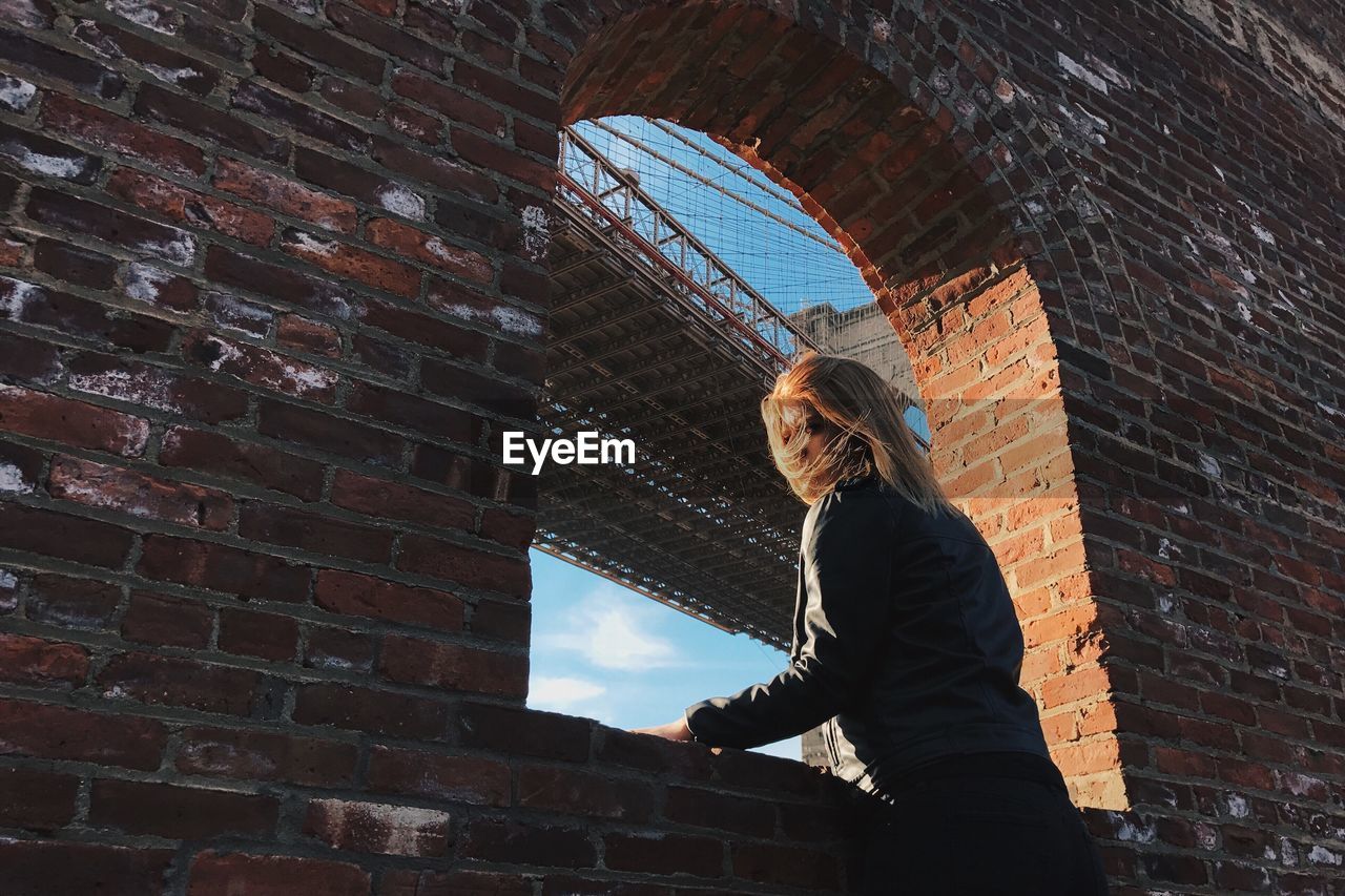 Low angle view of woman standing by brick wall against brooklyn bridge