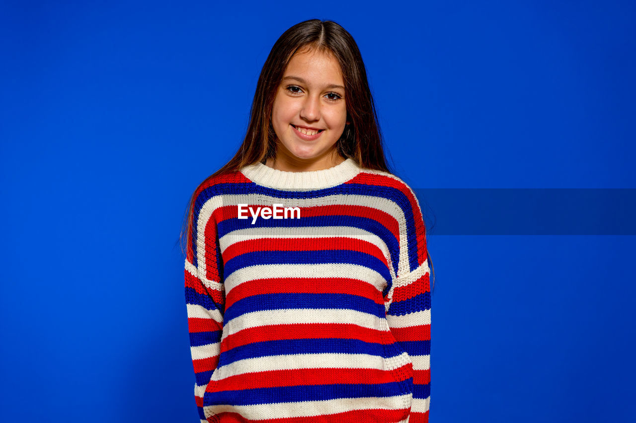 striped, blue, smiling, one person, happiness, portrait, studio shot, looking at camera, emotion, colored background, waist up, blue background, women, casual clothing, copy space, standing, adult, young adult, cheerful, hairstyle, front view, indoors, sweater, long hair, teeth, smile, photo shoot, clothing, positive emotion, brown hair, person, child, sleeve, fun, joy
