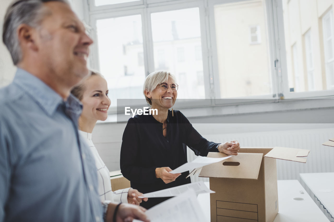 Smiling mature businessman with colleague unpacking cardboard box in new office