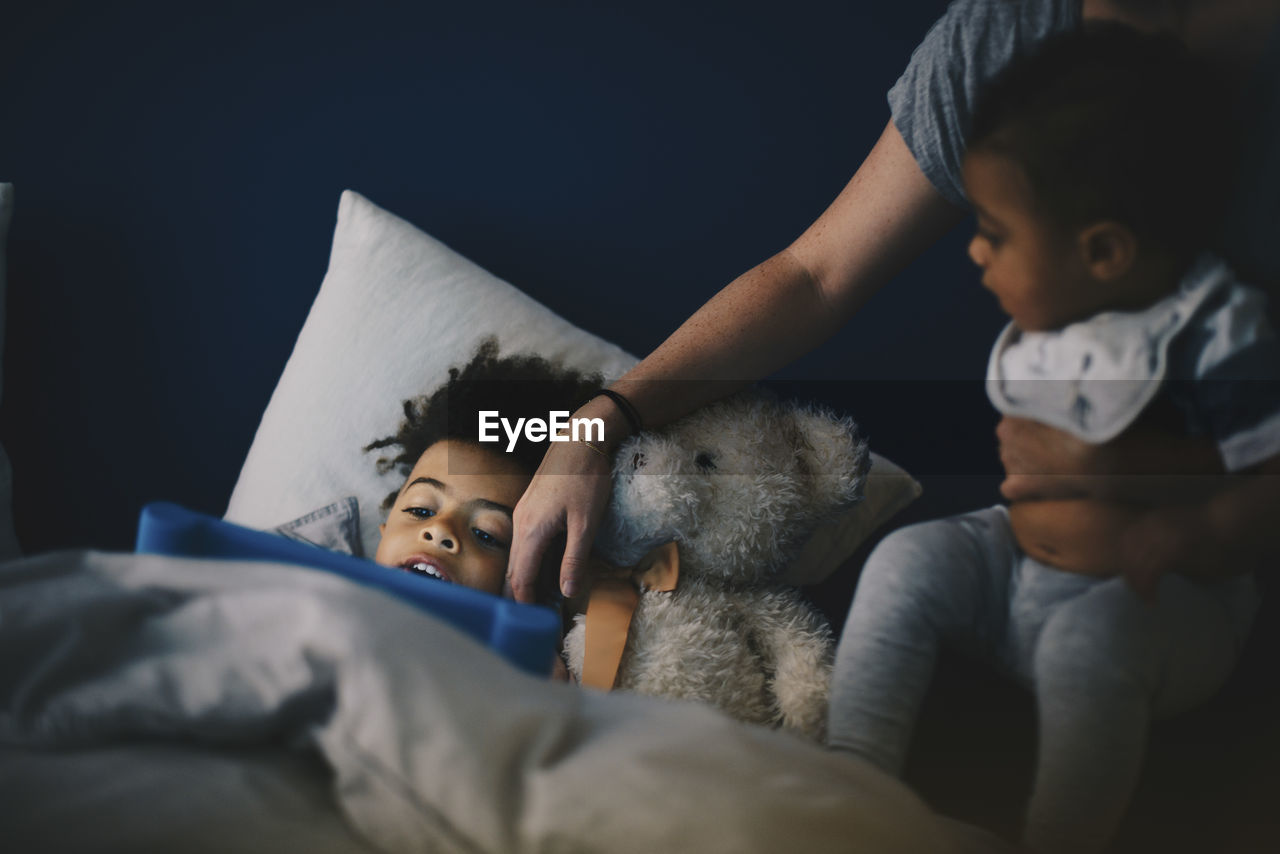 Mother carrying toddler sitting besides son using digital tablet on bed at home