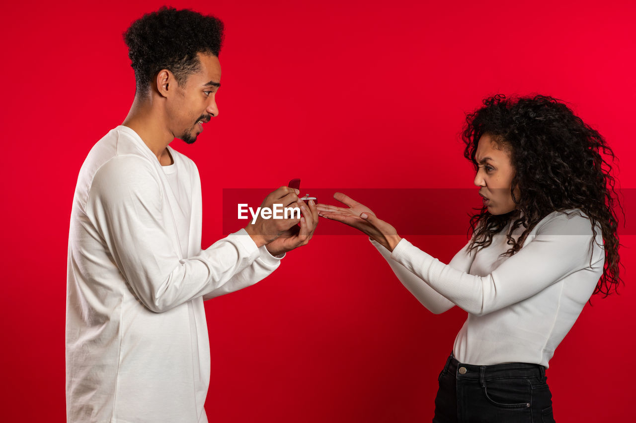 Woman making face while looking at ring held by boyfriend against red background