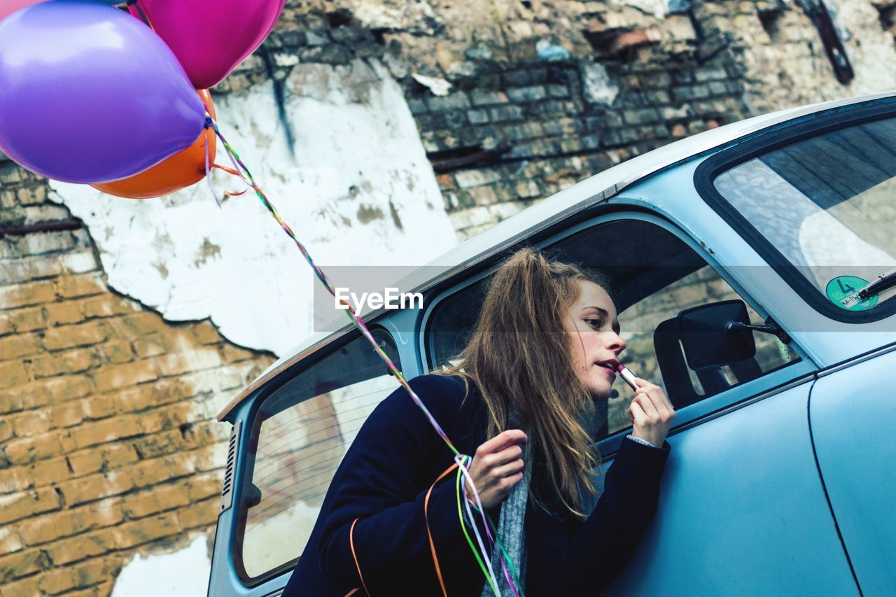 Young woman with colorful balloons applying lipstick while looking into side-view mirror of car