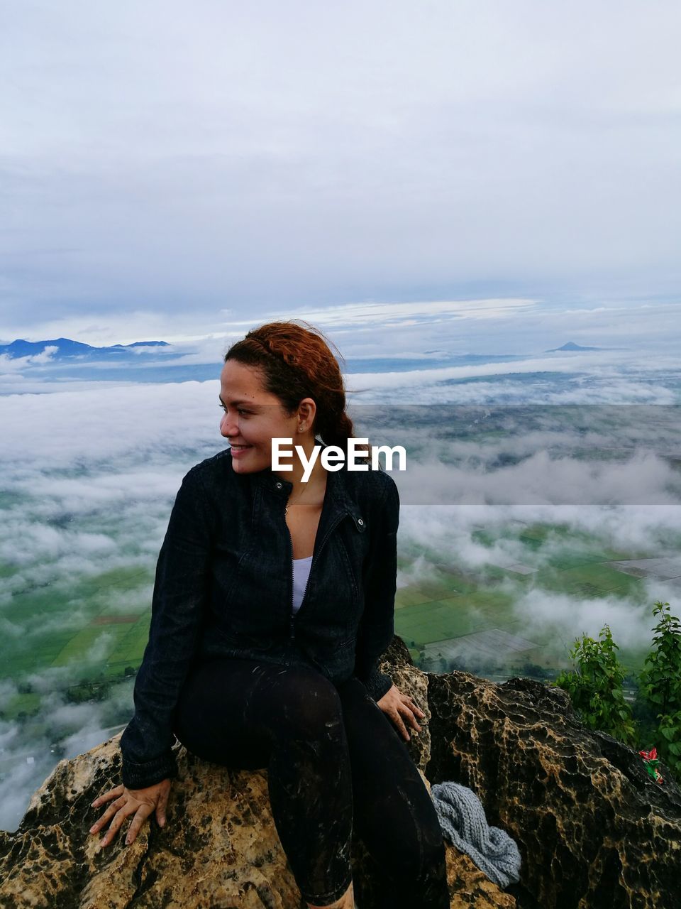 Smiling woman sitting on rock at mountain over clouds against sky