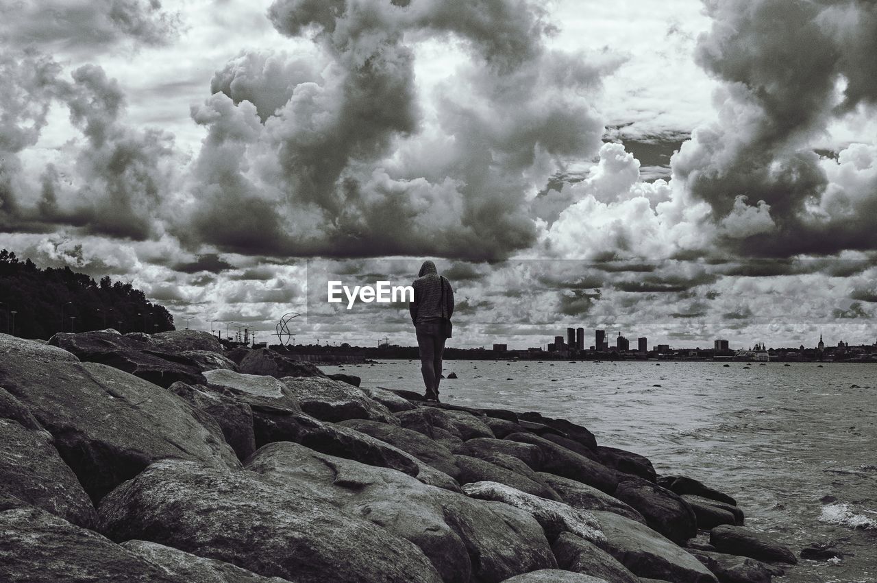 Rear view of man standing on rocks by sea in city against cloudy sky