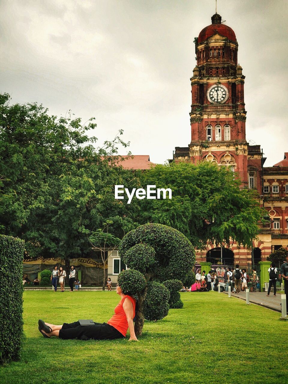 Woman sitting in garden by clock tower in city against cloudy sky