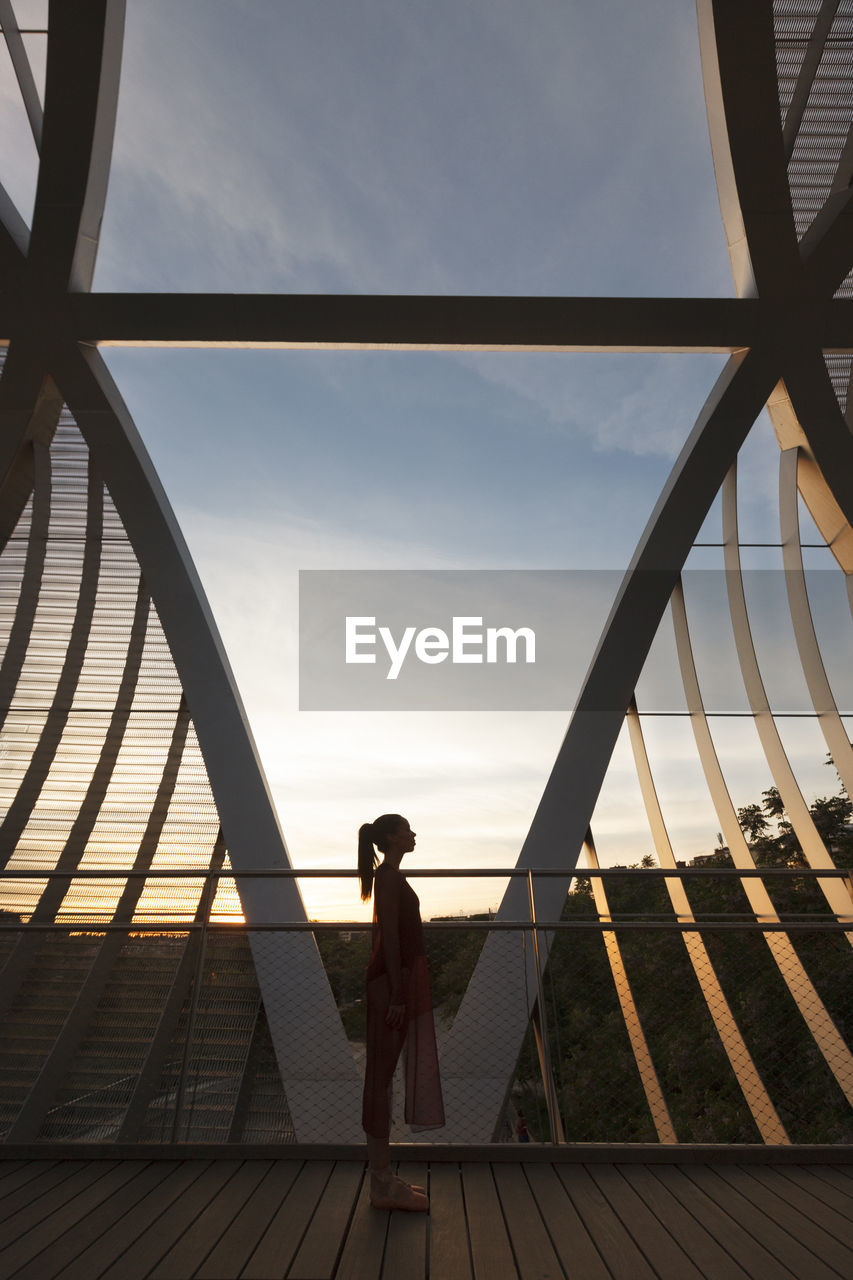 Side view of woman standing on footbridge against sky at sunset