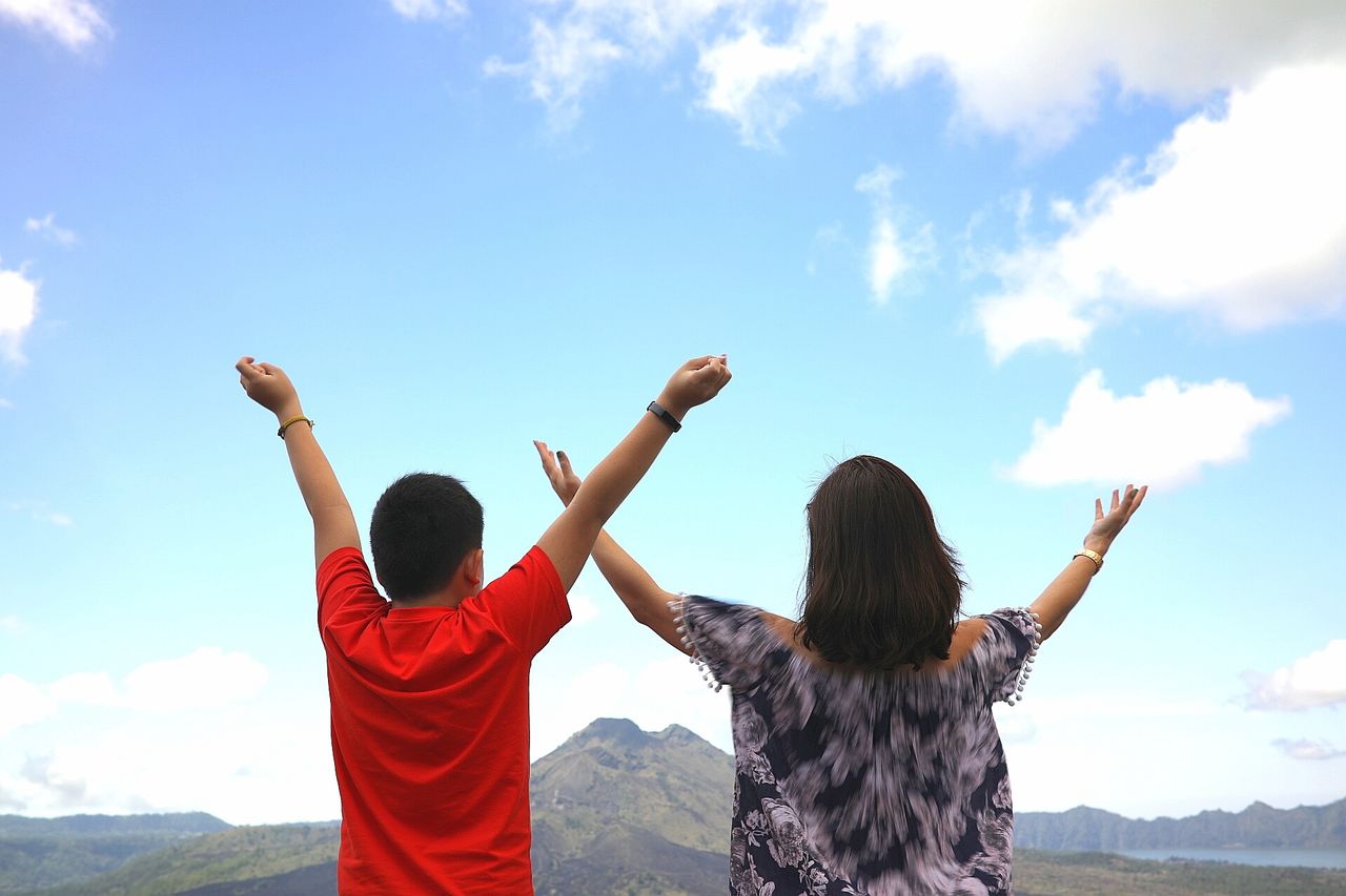 Rear view of mother and son with arms raised standing against sky
