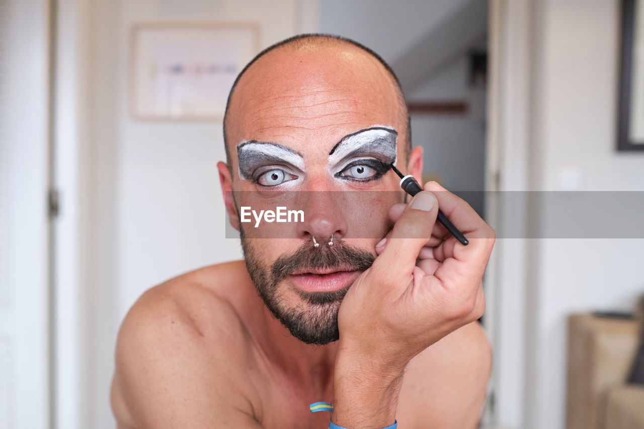 Adult shirtless bearded male transgender with eyebrow makeup and cosmetic lens applying black eyeliner