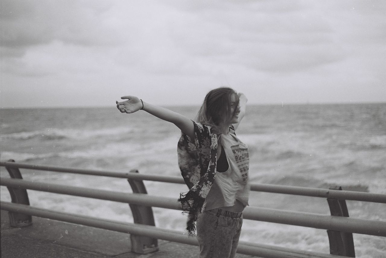 Side view of woman with arms outstretched standing by railing against sea