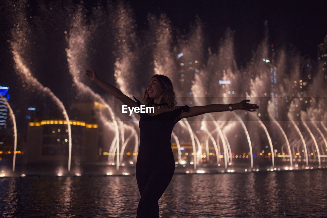 Woman with arms outstretched against fountain in city at night