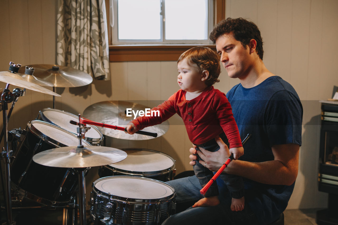 Father holding cute baby son playing drums at home