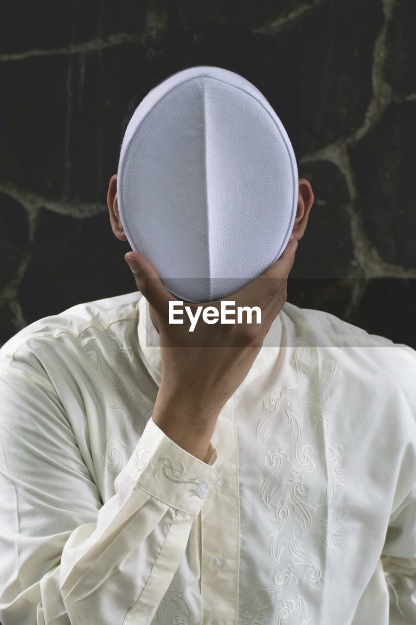 A fine art portrait of a man in indonesian islamic shirt hide his face with the religious white cap.