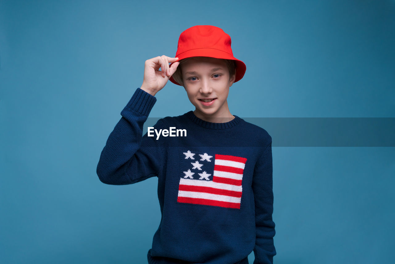 one person, studio shot, cap, red, portrait, colored background, blue, patriotism, t-shirt, hat, emotion, clothing, blue background, standing, men, looking at camera, indoors, waist up, baseball cap, child, front view, smiling, childhood, fashion accessory, pride, flag, young adult, adult, sleeve