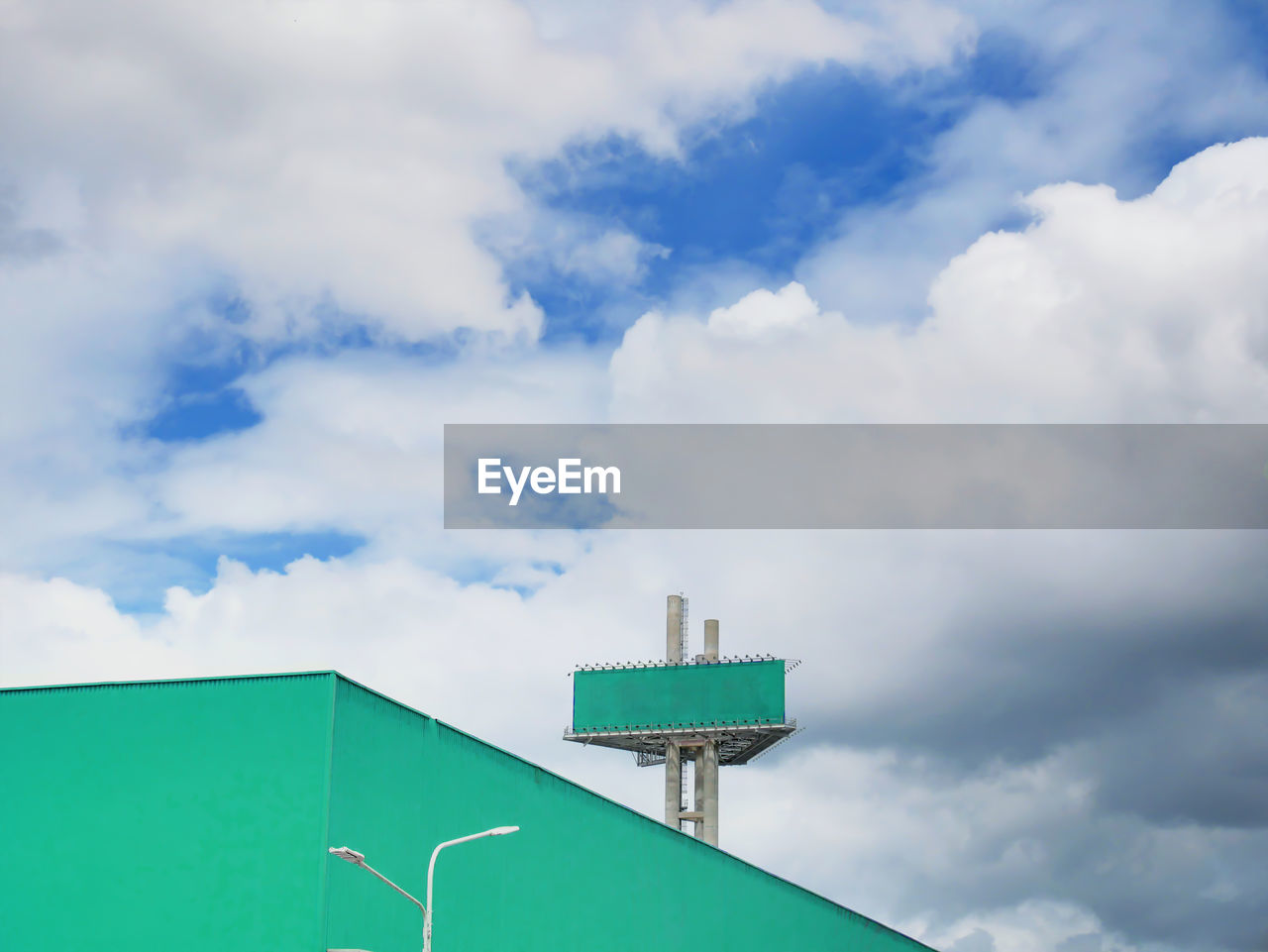 Green empty advertisement billboard on large green warehouse against blue cloudy sky