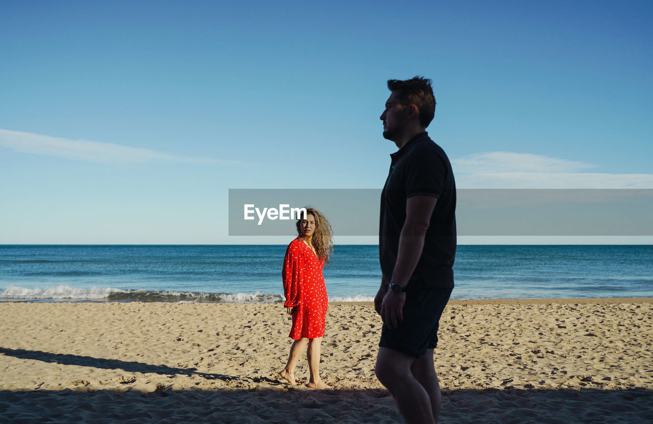 Rear view of woman standing standing behind man at beach against clear sky