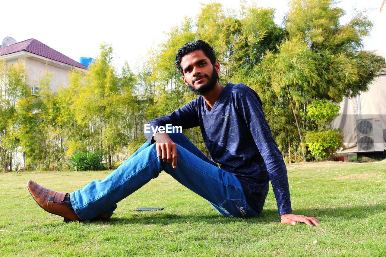 one person, plant, grass, men, young adult, adult, casual clothing, full length, smiling, nature, happiness, leisure activity, lifestyles, portrait, sitting, relaxation, tree, emotion, beard, day, facial hair, person, architecture, front view, green, looking at camera, jeans, outdoors, cheerful, clothing, fashion, building exterior, lawn, built structure, field, sunlight, park, looking, sky