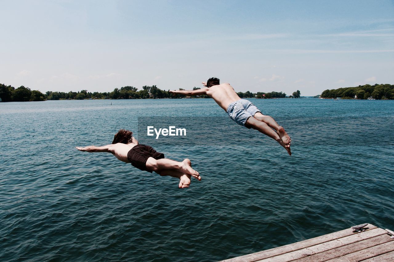 Shirtless friends jumping into river against sky