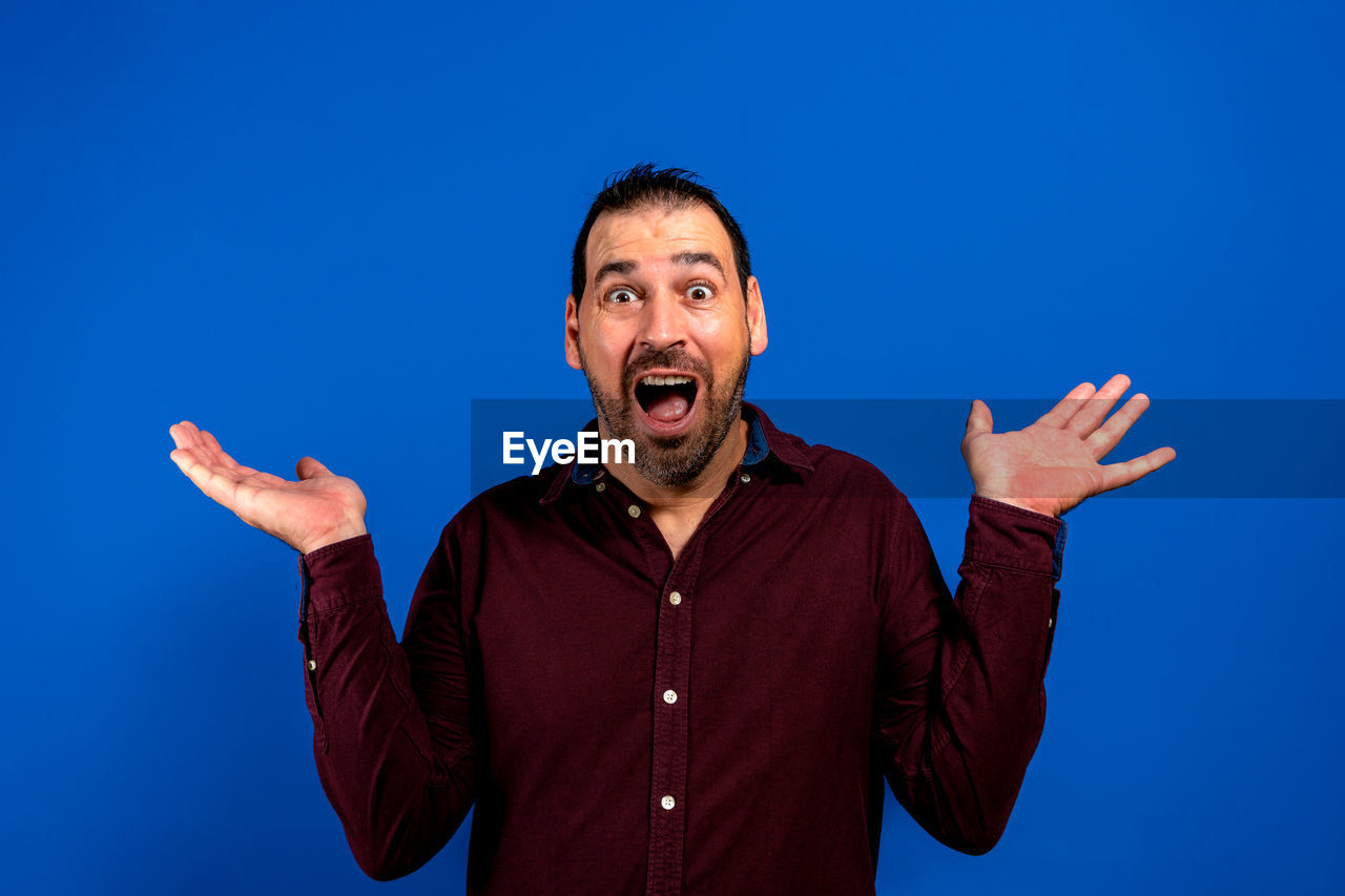blue, emotion, gesturing, one person, shouting, sign language, men, adult, studio shot, mouth open, happiness, portrait, waist up, positive emotion, colored background, person, excitement, cheerful, looking at camera, front view, hand, blue background, human mouth, singing, facial expression, smiling, fun, copy space, arm, anger, communication, limb, joy, surprise, young adult, human limb, negative emotion, casual clothing, indoors, furious, hand raised, hand sign, finger, ecstatic