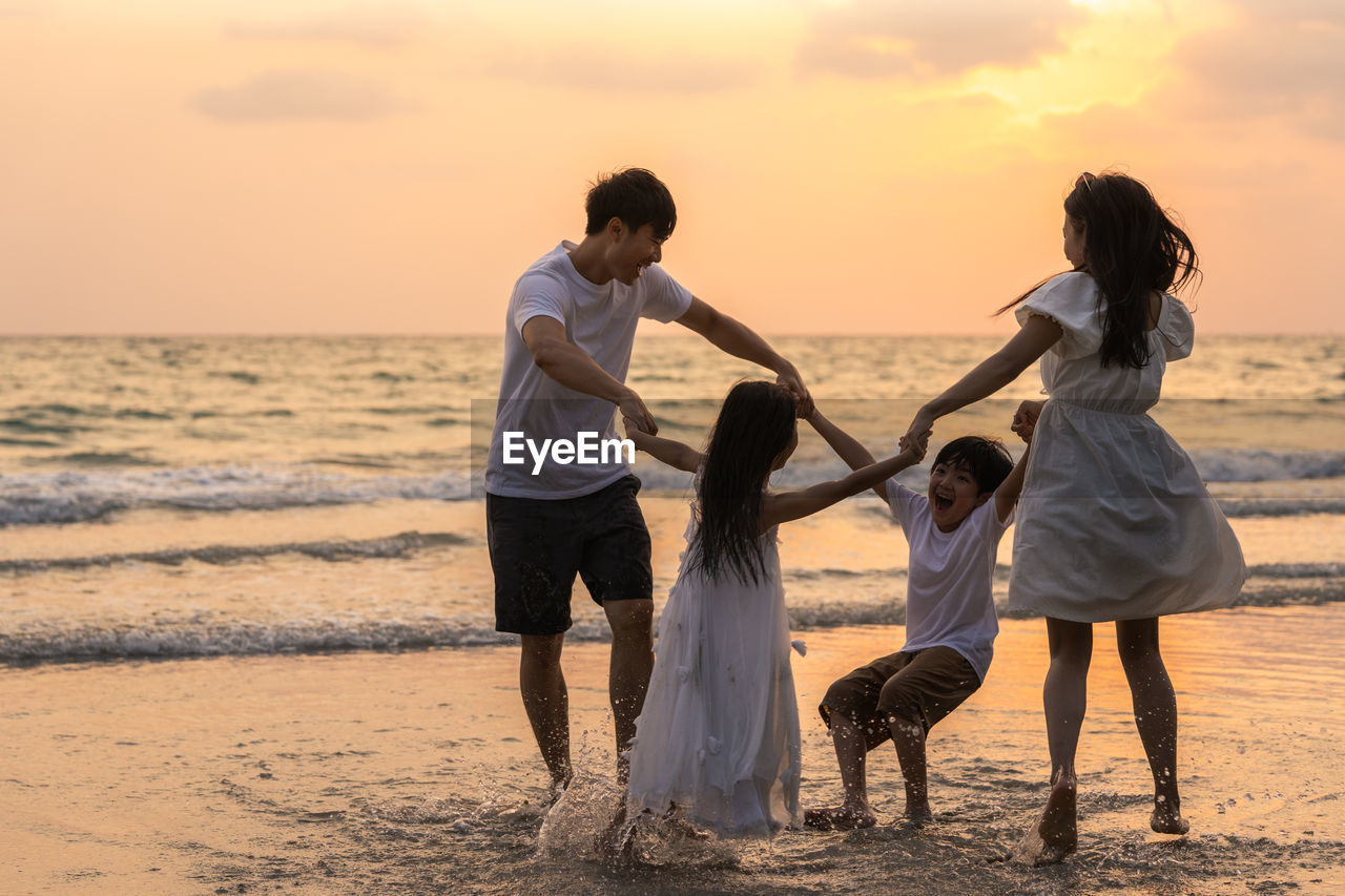 Family enjoying at beach against sky during sunset