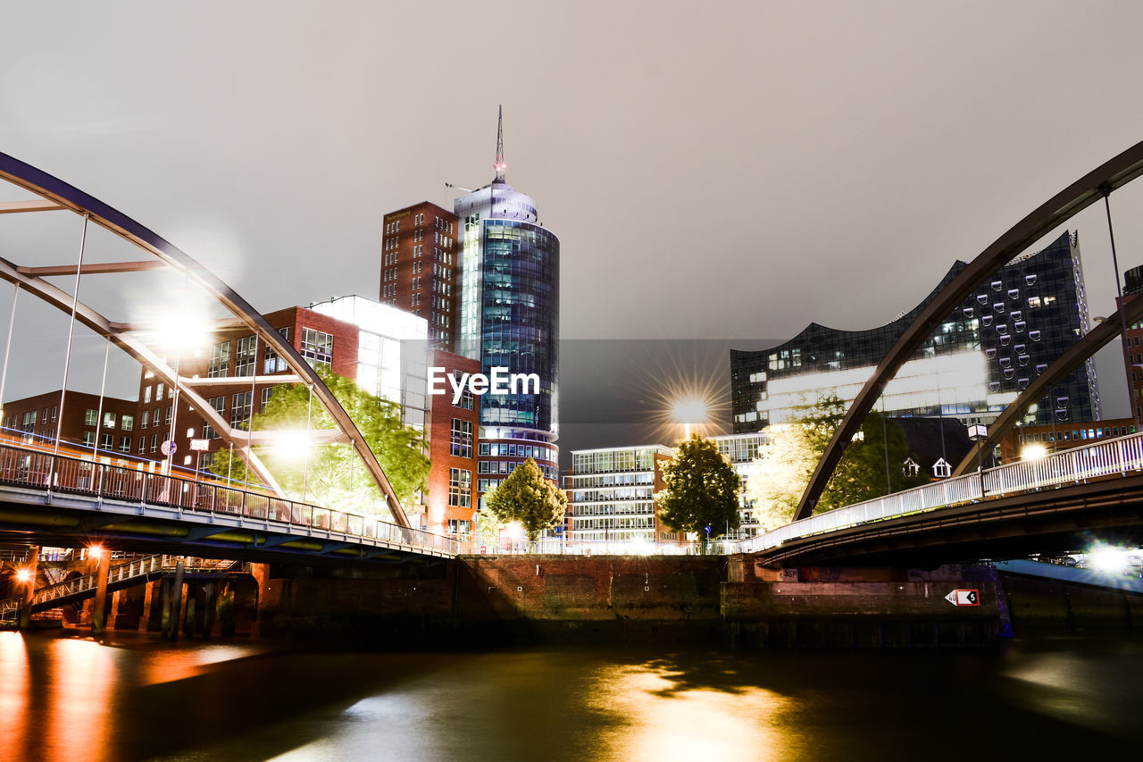 ILLUMINATED BRIDGE OVER RIVER BY BUILDINGS AGAINST SKY