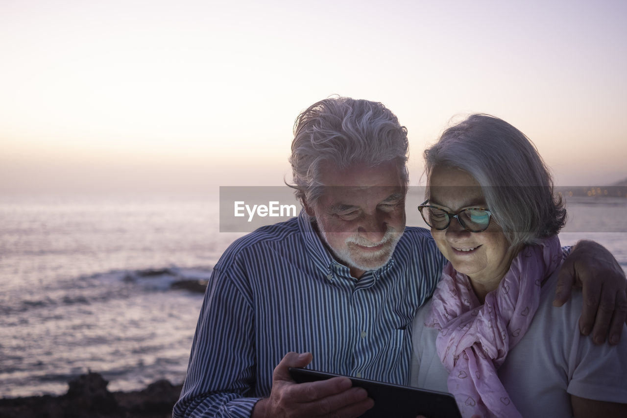 Friends looking at camera on beach against sky during sunset