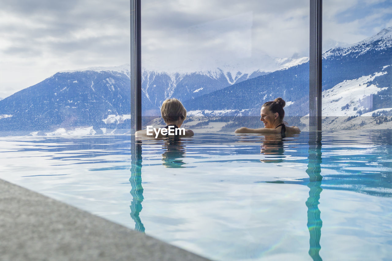 Rear view of female friends swimming in pool against snowcapped mountains