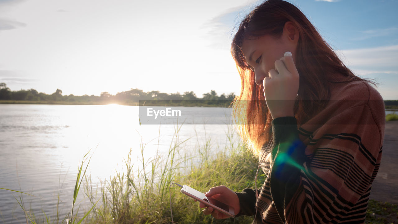 YOUNG WOMAN USING SMART PHONE AGAINST SKY DURING SUNSET