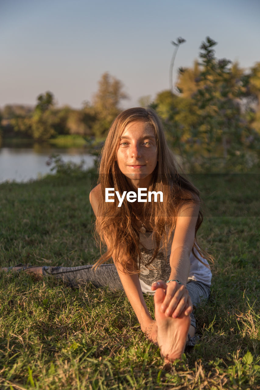 Portrait of teenage girl exercising on grass against sky during sunset