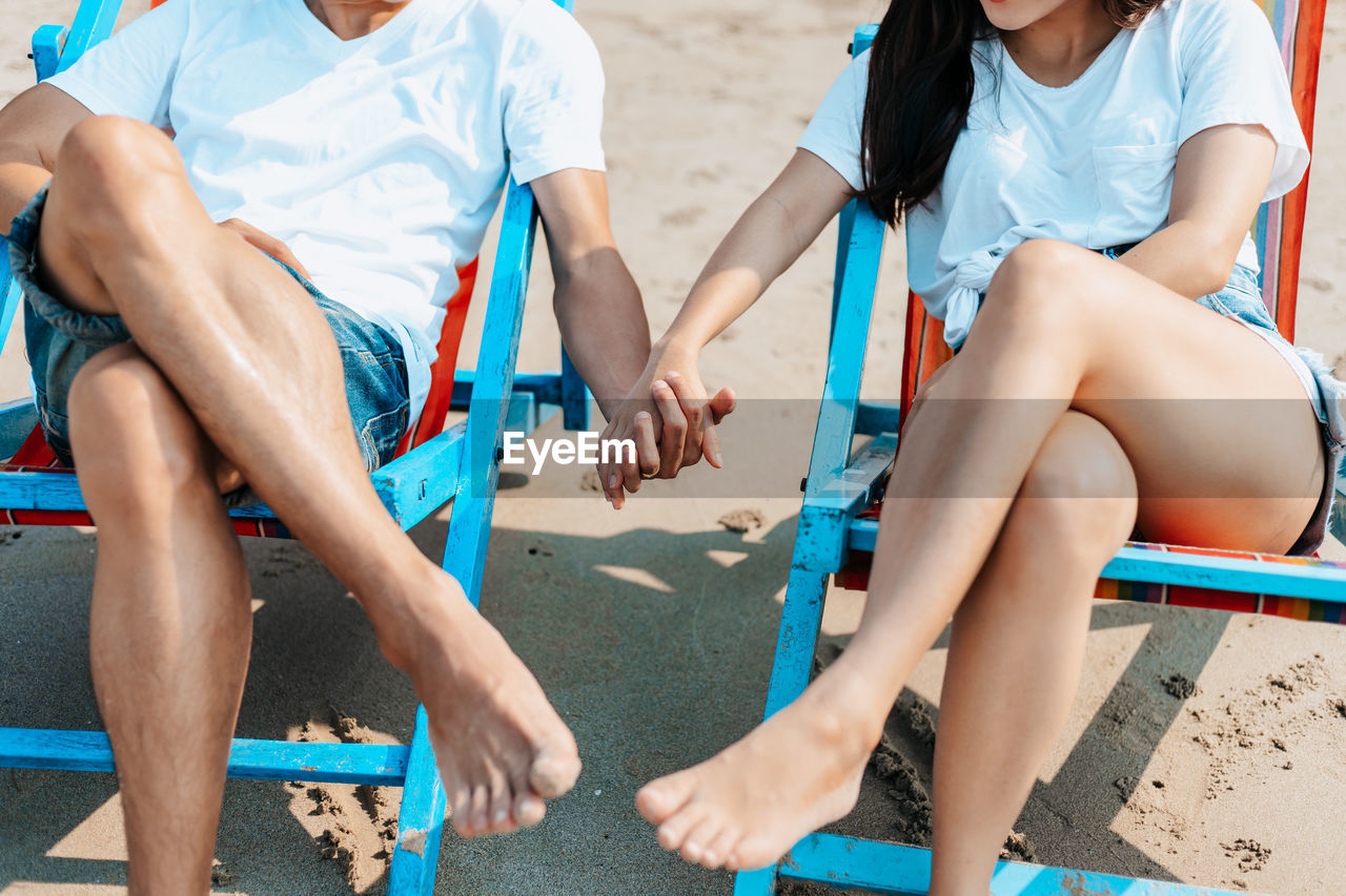 Low section of couple holding hands while sitting at beach