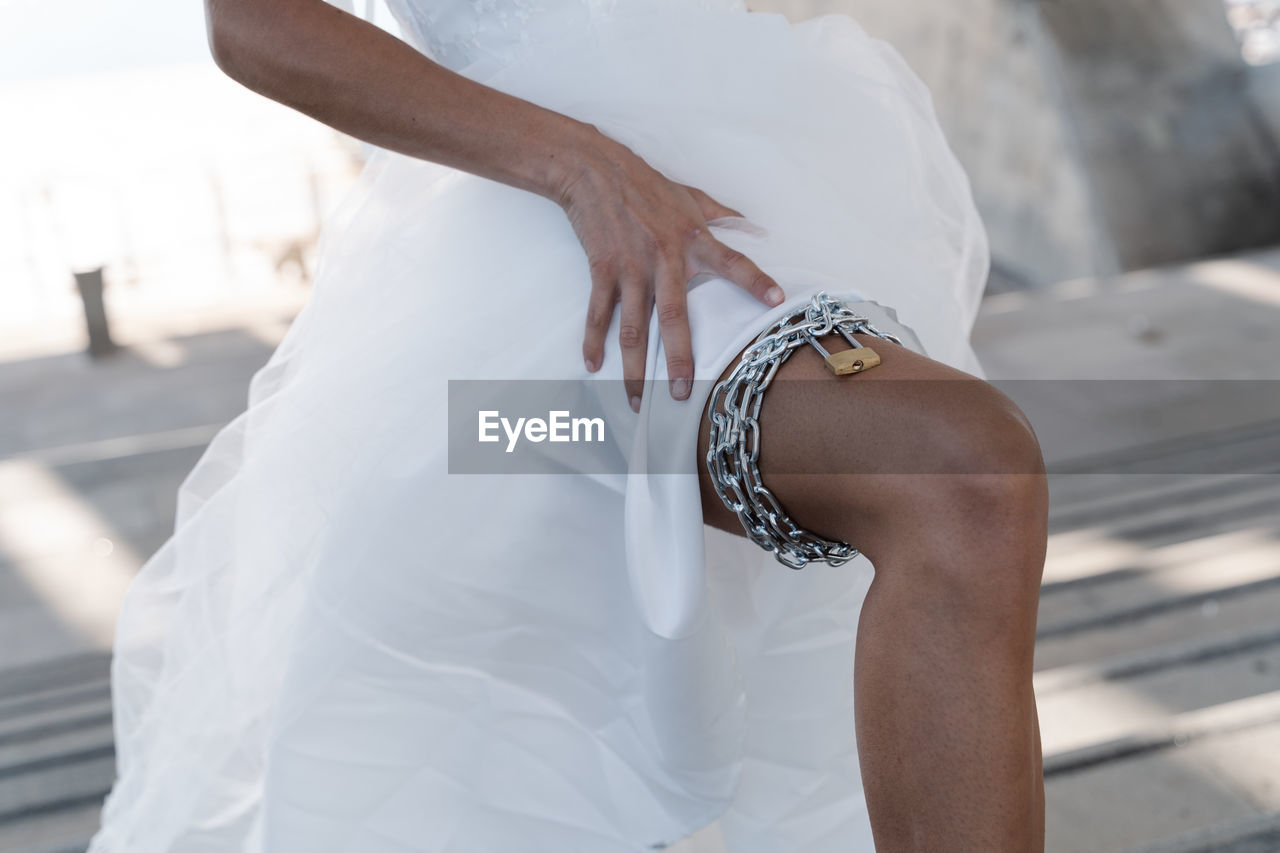 Crop unrecognizable female in elegant white wedding dress lifting leg showing chain garter
