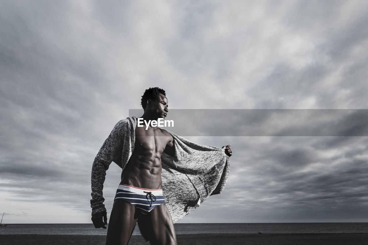 Low angle view of muscular man standing in underwear at beach against cloudy sky
