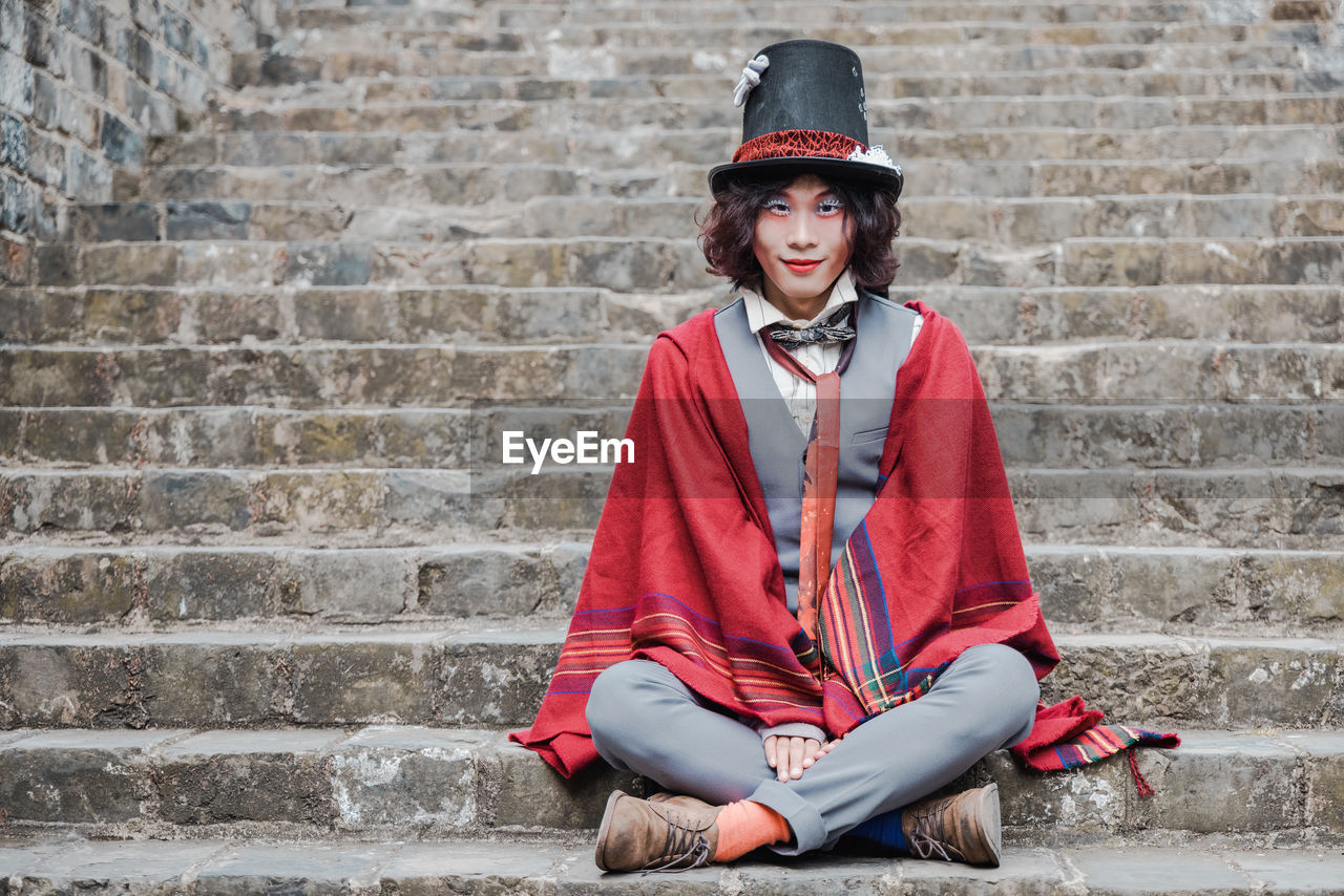 Portrait of man cosplaying mad hatter sitting on staircase against brick wall