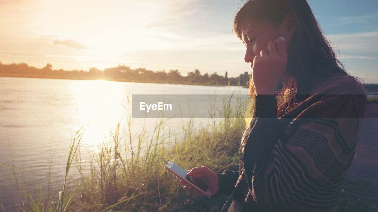 YOUNG WOMAN USING SMART PHONE BY LAKE AGAINST SKY