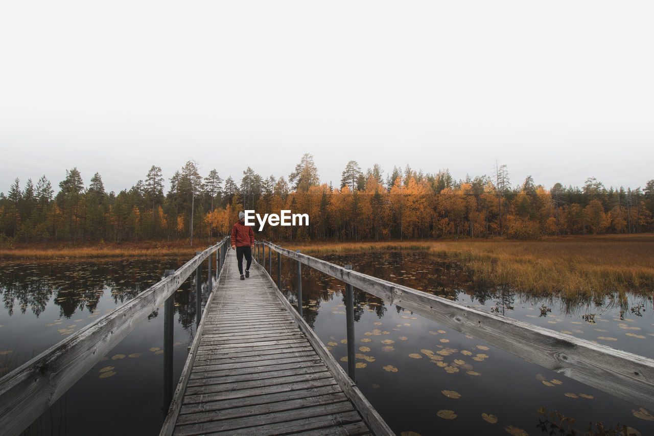 Man in a red jacket and cap walks on a wooden bridge, looking out over lake. kainuu region, finland.