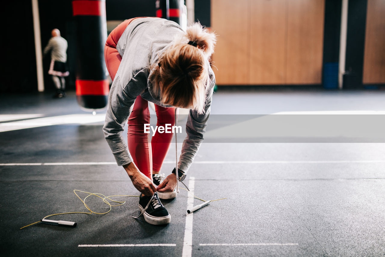 Unrecognizable female athlete in sportswear tying laces on sneakers while standing near hanging punching bag and preparing for workout in modern gym