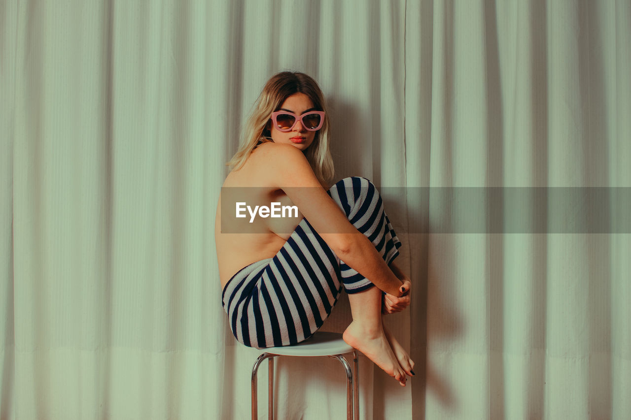 Side view of shirtless young woman wearing sunglasses while sitting on chair by curtain